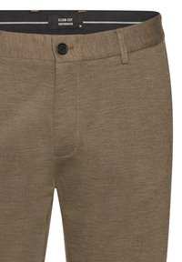 Pantalon marron ajusté en tissu texturé, avec une fermeture à bouton et un design épuré. Étiquette de la marque visible à la taille.