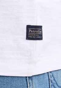 Bílá látka s texturovaným povrchem, na které se nachází černá etiketa se zlatým textem, který zní: "Petrol Ind Denim Driven Since '99 Florida, U.S.A."