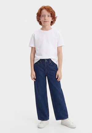 Enfant aux cheveux roux portant un t-shirt blanc uni, un jean large bleu foncé et des baskets blanches, debout devant un fond clair uni.