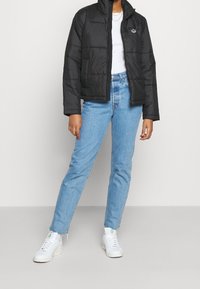 Czarna puchowa kurtka z wysokim kołnierzem i pikowanym wzorem, zestawiona z jasnoniebieskimi jeansami o prostych nogawkach i białymi tenisówkami.