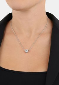 Silverkedja med ett hjärtformat hänge som pryds av en klar ädelsten. Halsbandet vilar mot en svart topp med en slät textur.