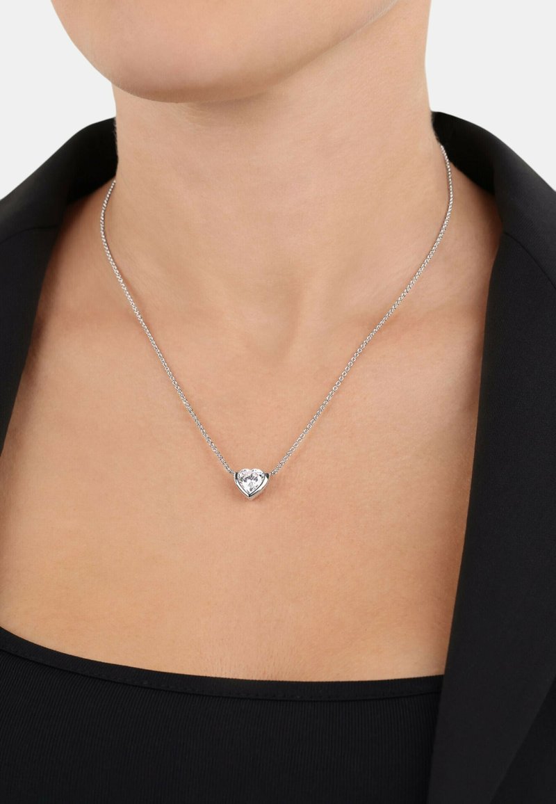Silverkedja med ett hjärtformat hänge som pryds av en klar ädelsten. Halsbandet vilar mot en svart topp med en slät textur.