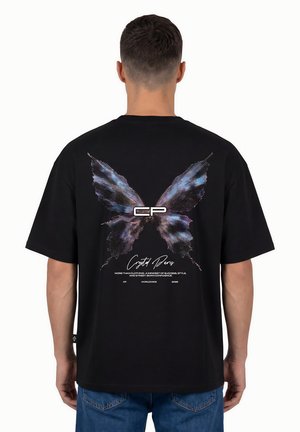 Homme portant un t-shirt noir avec un grand motif papillon bleu et violet avec "CP" et un texte au dos, assorti à un jean bleu.