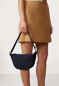 Borsa a tracolla in tessuto blu navy con forma curva, consistenza liscia e tracolla staccabile, tenuta da una mano sopra una gonna color beige.