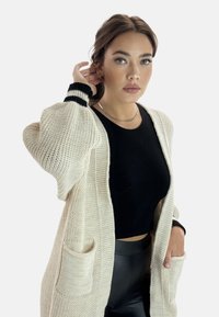 Lång, krämfärgad stickad cardigan med svarta detaljer. Har vida ärmar och två framfickor. Bärs över en svart topp och leggings.