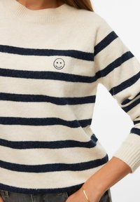 Gros plan sur un pull beige à rayures horizontales bleu foncé avec un petit visage souriant brodé sur la poitrine, porté par une personne portant un bracelet en or.