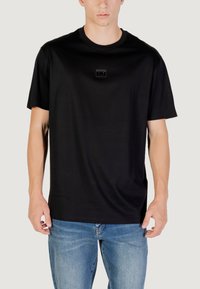 T-shirt nera a maniche corte in morbido tessuto con scollo rotondo e un'etichetta logo rettangolare sul petto. Indossata con jeans in denim blu.