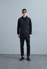 Fekete negyed-zippes pulóver, textúrált anyag, karcsú fazonú fekete nadrág és fekete cipő; a modell napszemüveget visel, egységes szürke háttér előtt.