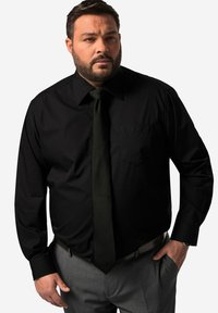 Una camicia nera a maniche lunghe con colletto a punta e taschino sul petto, abbinata a una cravatta verde scuro e pantaloni grigi, presenta una superficie liscia e una vestibilità sartoriale.