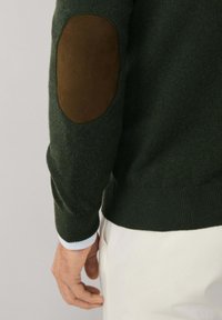Maglione verde lavorato a maglia con una toppa in suede marrone rotonda sui gomiti, polsini e vita a costine. La manica arriva fino al polso.