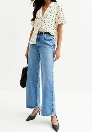 Femme portant un chemisier blanc à manches courtes avec des motifs d'étoiles noires, un jean bleu taille haute à jambes larges, des talons noirs et tenant un sac à main noir.