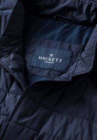 Marineblaue gesteppte Jacke mit hohem Kragen und einem Stoffetikett mit der Aufschrift "HACKETT LONDON" in weißer Schrift, glatte Textur.