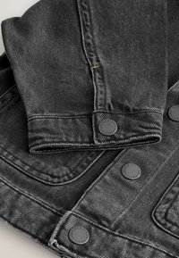 Gros plan des manches d'une veste en jean noire et des boutons métalliques avec des coutures piquées et un détail de fil jaune.