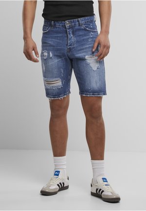 Mann iført slitte blå denimshorts, hvite sokker og hvite joggesko med svarte striper, står mot en ensfarget grå bakgrunn.