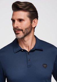 Polo shirt navy con colletto a punta, patta con tre bottoni e un'etichetta logo triangolare sul lato sinistro del petto. Texture liscia.