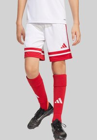 Rote und weiße Fußballshorts mit drei roten Streifen und Adidas-Logo. Rote Kniesocken mit weißem Logo. Schwarze Fußballschuhe mit strukturiertem Design.