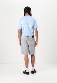 Lyseblå bomulds t-shirt med teksten "LES CLASSIQUES", parret med grå denimshorts. Individet bærer sorte sandaler og hvide sokker.