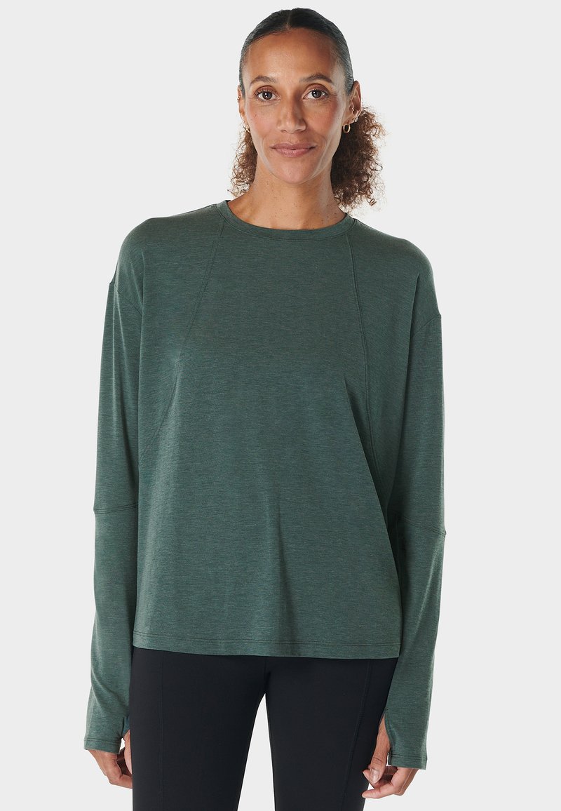Vrouw met teruggehaald krullend haar, gekleed in een groene lange mouwen shirt en zwarte broek, staand tegen een effen lichte achtergrond.