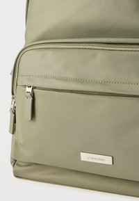 Μπροστινή τσέπη σακιδίου πλάτης από ανοιχτό πράσινο ύφασμα με δύο φερμουάρ και μια μικρή μεταλλική πλακέτα λογότυπου Calvin Klein τοποθετημένη στο κέντρο κάτω από την τσέπη.