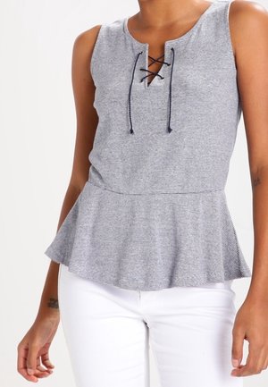 Mouwloze grijs-wit gestreepte peplum top met veterdetail bij de hals, gedragen met witte broek.