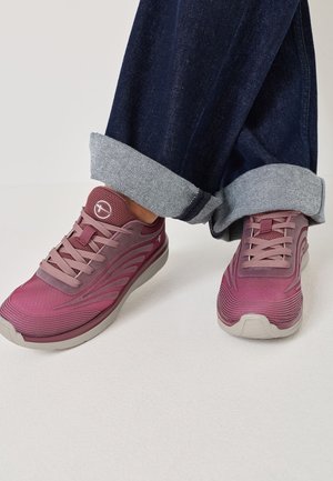 Lilla atletiske sneakers med lyserøde snørebånd båret med opkrøllede mørkeblå jeans på en hvid gulv.