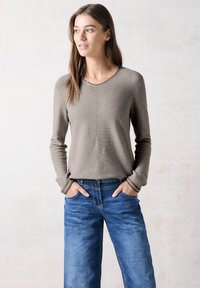 Top a maniche lunghe color marrone chiaro a coste con scollo a V; abbinato a jeans blu larghi. Il top presenta un design semplice e pulito con dettagli di cucitura.