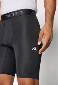 Mustana sportliku lõikega lühiülikond, millel on sujuv tekstuur ja "TECHFIT" sildiga vöö. Adidas logo on küljes valgena.