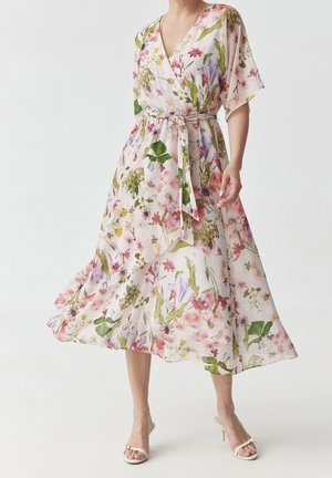 Femme portant une robe portefeuille fluide à fleurs avec une ceinture nouée et des manches courtes, associée à des sandales à talons blanches à bouts ouverts.