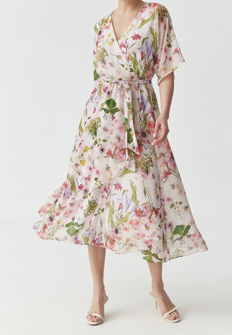 Femme portant une robe portefeuille fluide à fleurs avec une ceinture nouée et des manches courtes, associée à des sandales à talons blanches à bouts ouverts.