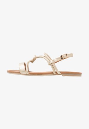 Złote metaliczne płaskie sandały z węzłowymi detalami, cienkimi paskami i regulowanym zapięciem na kostce. Płaska podeszwa, minimalistyczny design.