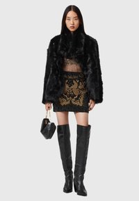 Syntetyczny czarny płaszcz futrzany, przezroczysta bluzka z ozdobami, złota i czarna mini spódnica z koralikami, wysokie czarne skórzane botki, trzymając małą czarną torebkę.