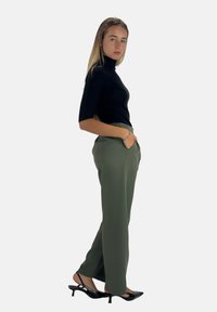 Maglione nero aderente a collo alto abbinato a pantaloni verde oliva con vita elastica e sandali neri con tacco. La modella è in posizione di profilo.