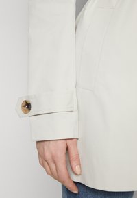 Chaqueta beige claro con una textura suave, que presenta un puño ancho asegurado por un botón redondo y translúcido. Mano reposando sobre la tela de la chaqueta.