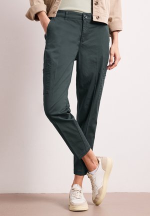 Personne portant un pantalon cargo court vert foncé et une veste beige aux manches retroussées, associée à des baskets blanches et beiges, debout les jambes croisées.