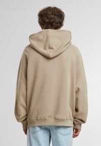 Beige kapucnis pulóver, puha textúrával, esésekkel a vállaknál és bordás mandzsettákkal és aljjal. Világoskék farmernadrággal viselve.