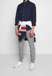 Marinblå skjorta med knappar, grå sweatpants, vita sneakers och en flerfärgad tröja knuten i midjan. Mjuk bomullstextur, avslappnad design.