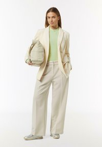 Beige pinstripe blazer over een lichtgroene top, brede crèmekleurige broek en zilveren sneakers. Ze houdt een luchtige, lichtgroene tas vast.