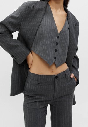 Femme portant un costume gris foncé à fines rayures avec un gilet court boutonné, un pantalon assorti et un blazer oversize posé sur les épaules.