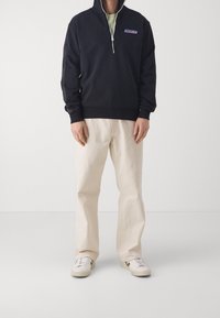 Sudadera negra con cremallera y cuello alto, con un logo en el pecho, combinada con pantalones de pierna ancha color crema y zapatillas blancas.