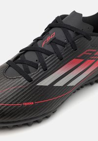 Svart fotbollssko med horisontell ribbad textur, röda och silverfärgade detaljer samt svarta skosnören. Innehåller "F50"-märke och "FIBERSKIN"-detalj.
