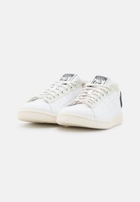 Bílé kožené tenisky s perforovanými detaily, plochými tkaničkami a gumovou podrážkou s kontrastním černým patním prvkem a logem.