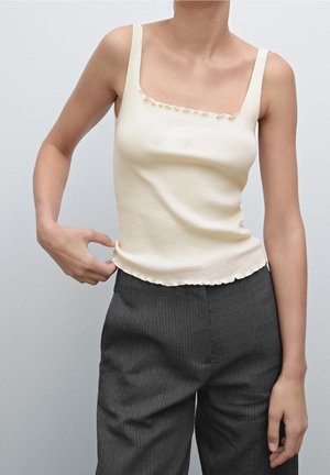 Femme portant un débardeur côtelé crème avec des bords froncés et un pantalon gris foncé à fines rayures, ajustant l'ourlet supérieur avec la main gauche.