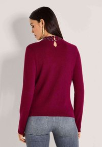Eine Frau mit langen, dunklen Haaren trägt einen enganliegenden, dunkelroten Pullover mit Knopfdetails am hinteren Kragen, kombiniert mit grauen Jeans.