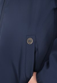 Mørkeblå jakke med en glat overflade, der har et metal logo mærke formet som en anker på venstre ærme. Lynlåsen er fremtrædende.