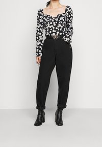 Kvinna som bär en svart blommig långärmad topp, högmidjade svarta byxor med bälte och svarta ankelskor, stående mot en enfärgad bakgrund.