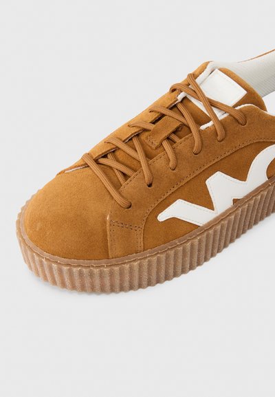 Καφέ αθλητικό παπούτσι από σουέτ με στρογγυλεμένη μύτη, λευκό λογότυπο και υφή στη σόλα από καουτσούκ. Διαθέτει επίπεδο σύστημα κορδονιών και επενδεδυμένο αστράγαλο για υποστήριξη.