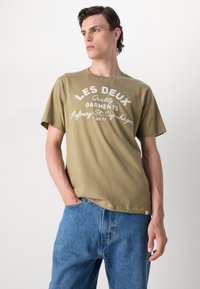 Olivovorosé tričko s bielym grafickým nápisom "LES DEUX Quality Garments" a uvoľneným strihom, skombinované so svetlomodrými džínsovými šortkami.