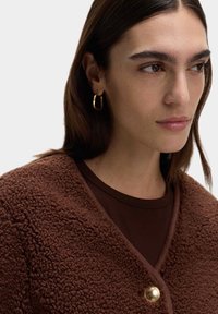 Giacca di pile marrone con una finitura testurizzata, caratterizzata da un dettaglio con bottone dorato. Indossata sopra una maglietta marrone scuro. Orecchino a cerchio dorato visibile.
