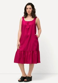 Fuchsiové midi šaty s volánkovými ramínky, rozšířeným lemem s volánky a bočními kapsami. Modelka má na sobě černé tkané sandály.