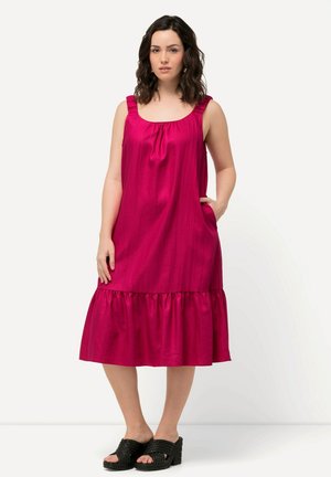Robe midi fuchsia avec des bretelles à volants, un ourlet à volants évasé et des poches latérales. Modèle portant des sandales tressées noires.